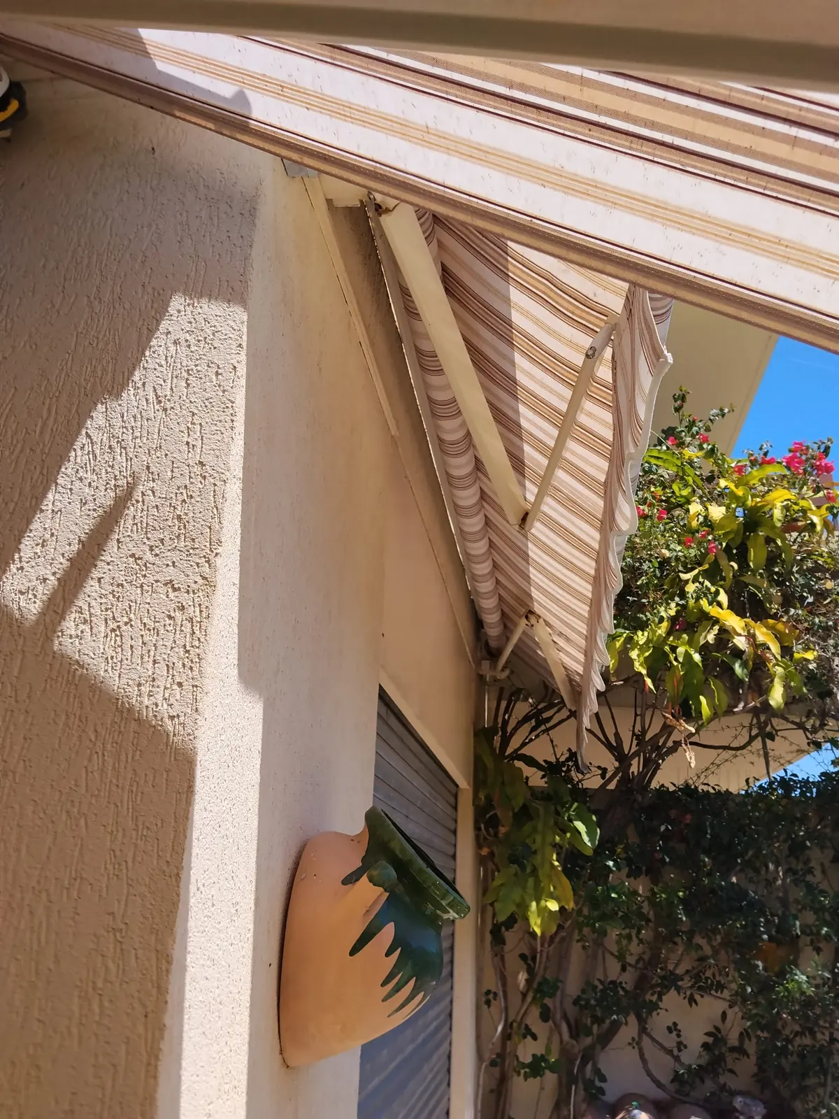 Store banne à rayures beige et brunes au-dessus d’une terrasse bordée de végétation.